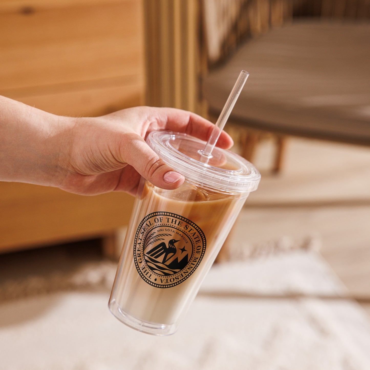Hand holding a clear cup with a straw, featuring the Minnesota State Seal, against a blurred indoor background.