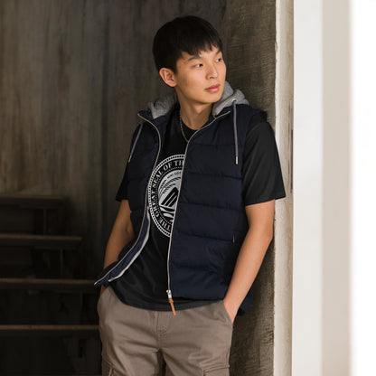 Person wearing a navy puffer vest over a black shirt featuring the Minnesota State Seal, standing against a neutral wall.